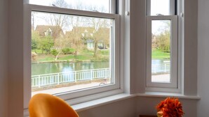A room with a window overlooking a body of water and a park, a chair, and a vase with flowers. - Riverside GEM 6BR With River View NR City Centre (Cambridge)
