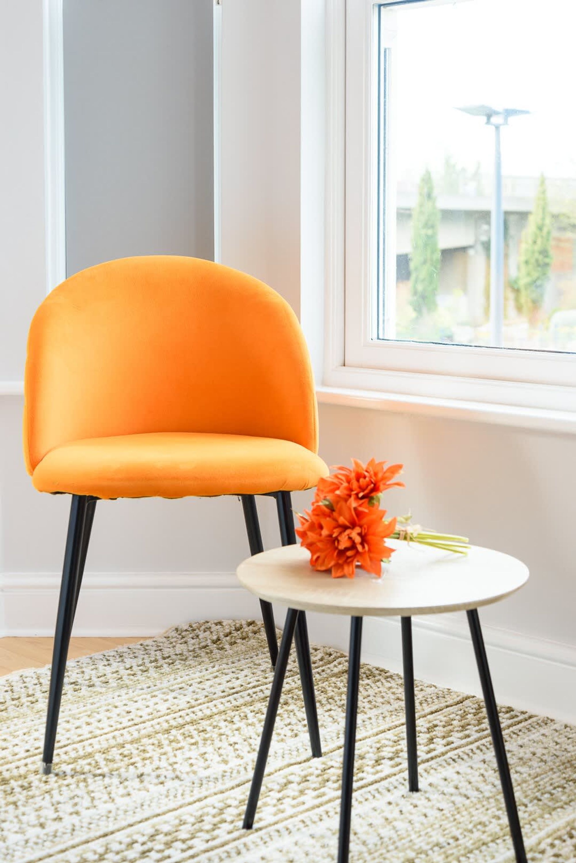 Una habitación con una única silla naranja, una pequeña mesa blanca con un ramo de flores naranjas, y una ventana que ofrece vistas a árboles y una calle.