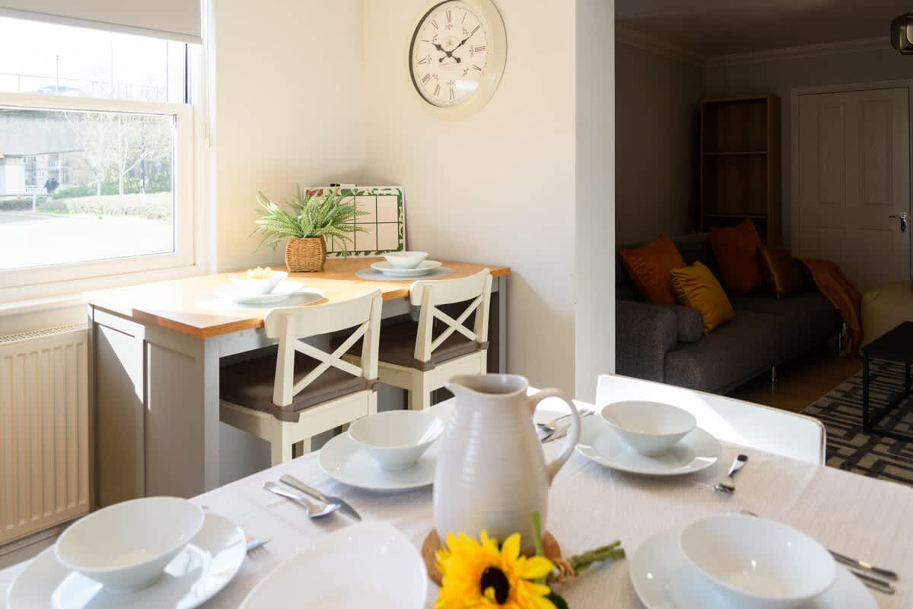 A dining area with a table set for two, a clock on the wall, and a sofa in the background.