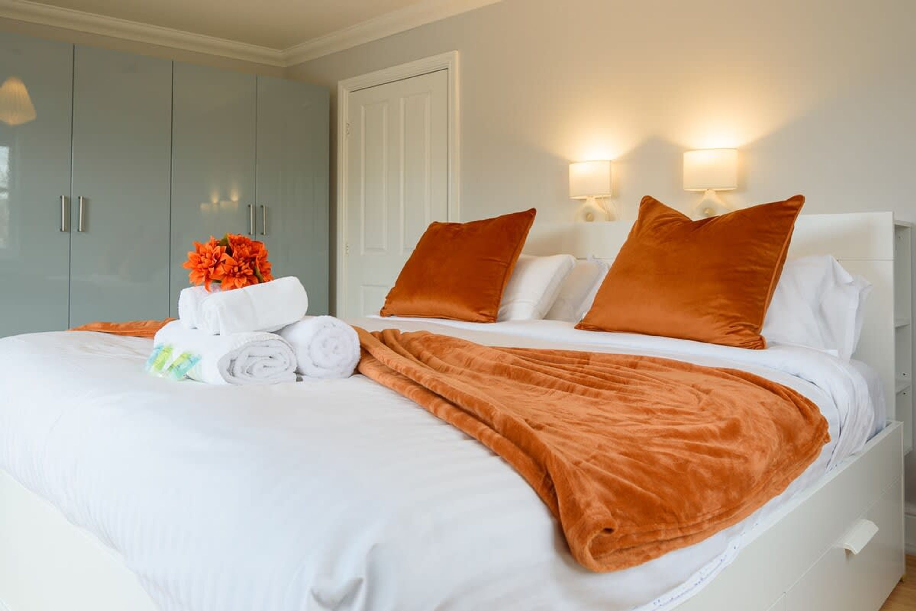 Cama bem arrumada com lençóis brancos e almofadas laranjas, um conjunto de toalhas e um arranjo decorativo de flores laranjas.