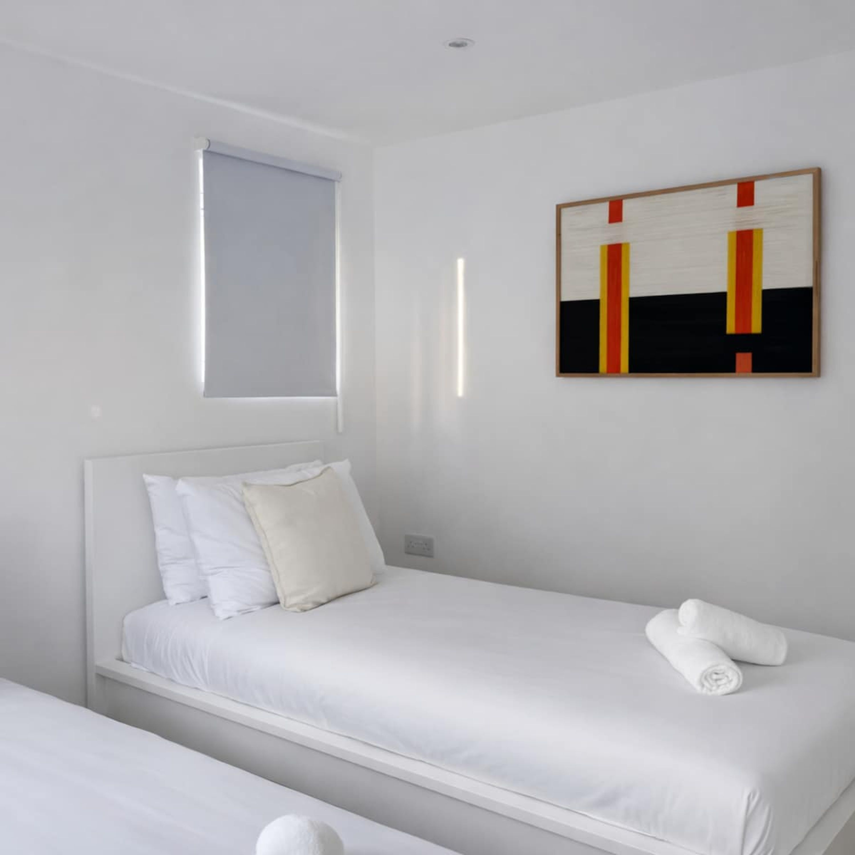 Ein schlicht eingerichtetes Hotelzimmer mit Bett, Kissen, einem Fenster und einem an der Wand befestigten Kunstwerk.