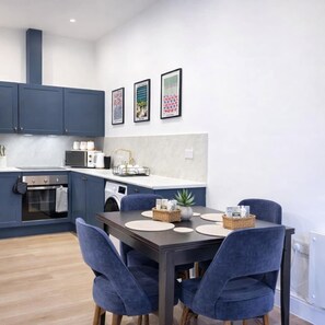 A modern kitchen with dark blue cabinets, a dining table with blue chairs, and framed artwork on the wall. - Dettori 2BR With Garden 11 Mins Walk to Gallops (Newmarket)