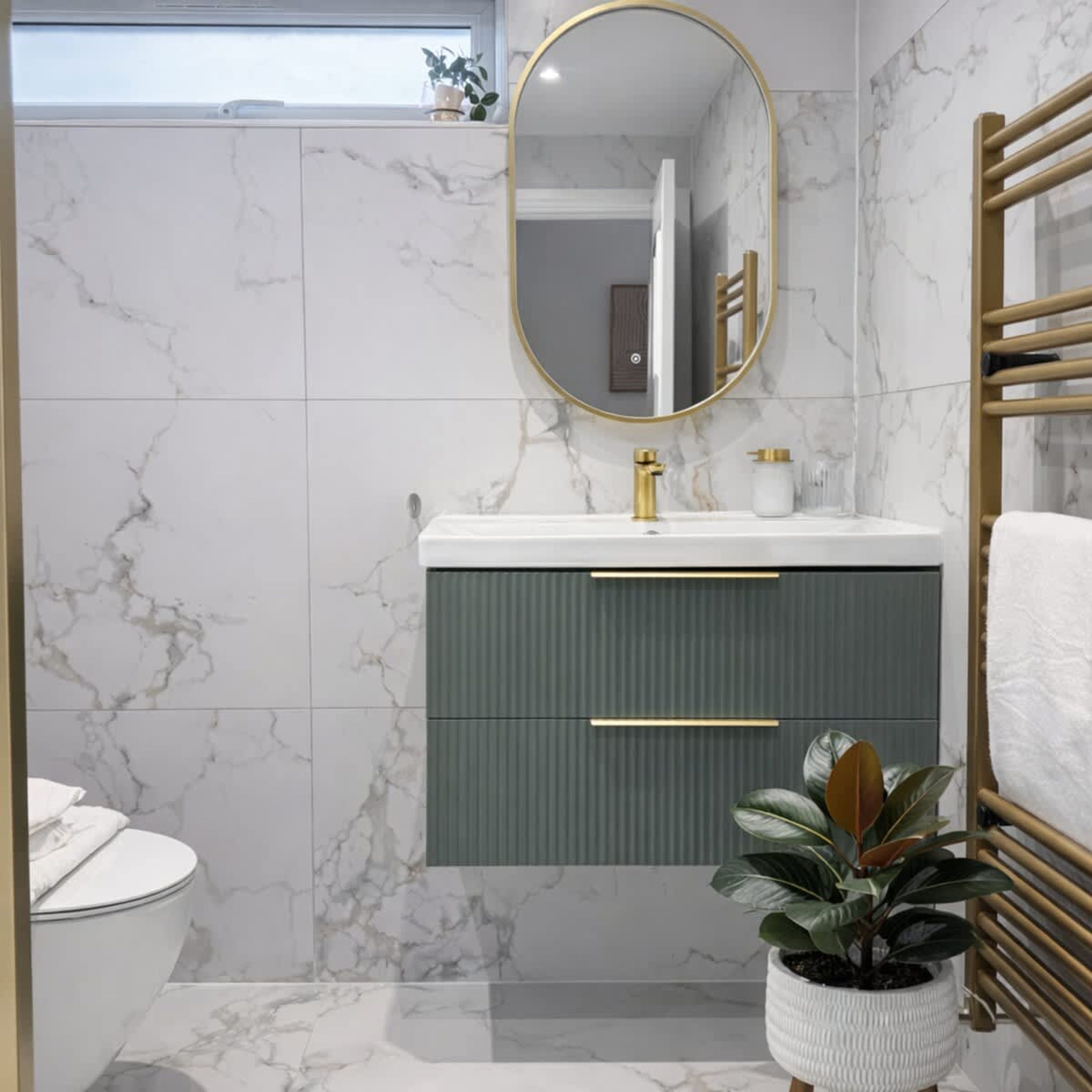 A bathroom with marble walls, a gold-framed mirror, a white sink with gold fixtures, a green cabinet, a white toilet, and a potted plant.
