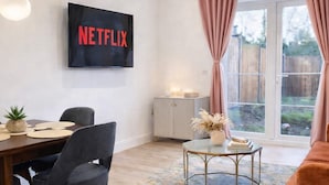 A modern living room with a dining area, a TV mounted on the wall, and a large window with curtains. - Dettori 2BR With Garden 11 Mins Walk to Gallops (Newmarket)