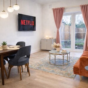 A modern living room with a dining area, a TV mounted on the wall, and a large window with curtains. - Dettori 2BR With Garden 11 Mins Walk to Gallops (Newmarket)