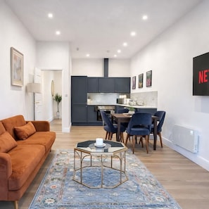 A modern living room with a sofa, a coffee table, and a dining area with chairs and a table. - Dettori 2BR With Garden 11 Mins Walk to Gallops (Newmarket)
