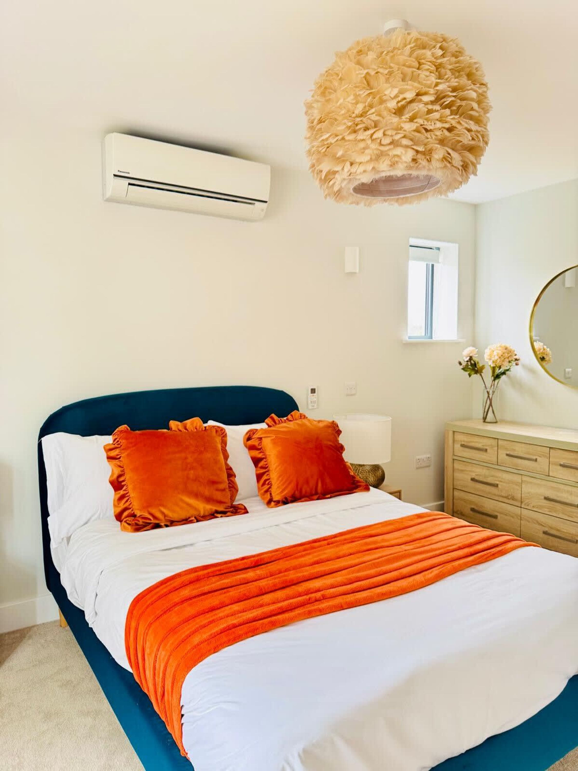 A bedroom with a bed, an air conditioning unit, a dresser, a mirror, and a hanging light fixture.