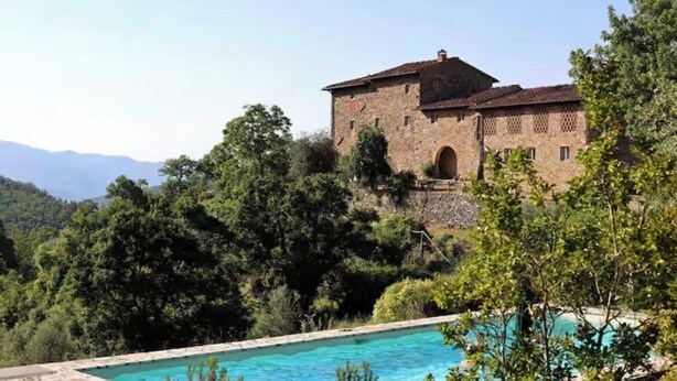 Um edifício de pedra com piscina, rodeado por árvores e colinas.