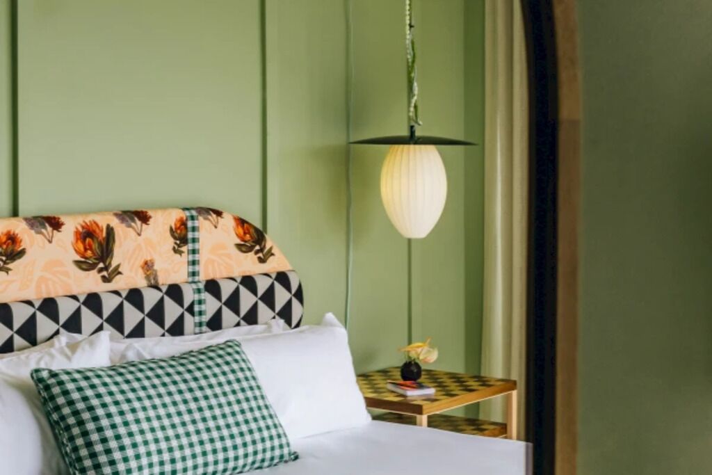 Uma cama com cabeceira estampada, um travesseiro verde xadrez e uma pequena mesa de cabeceira com um vaso de flores.