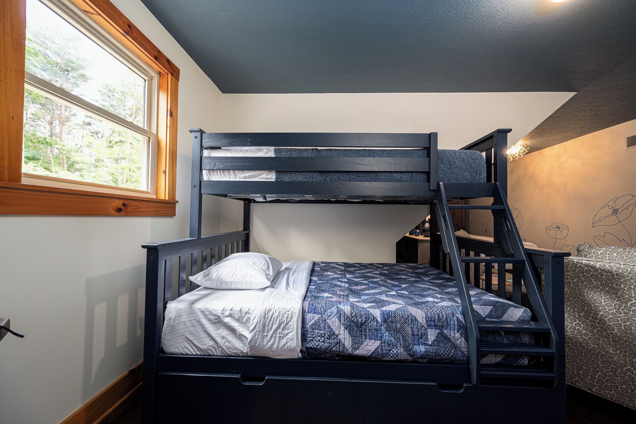 A bunk bed with a ladder, a window with a view of trees, and a patterned bedspread.