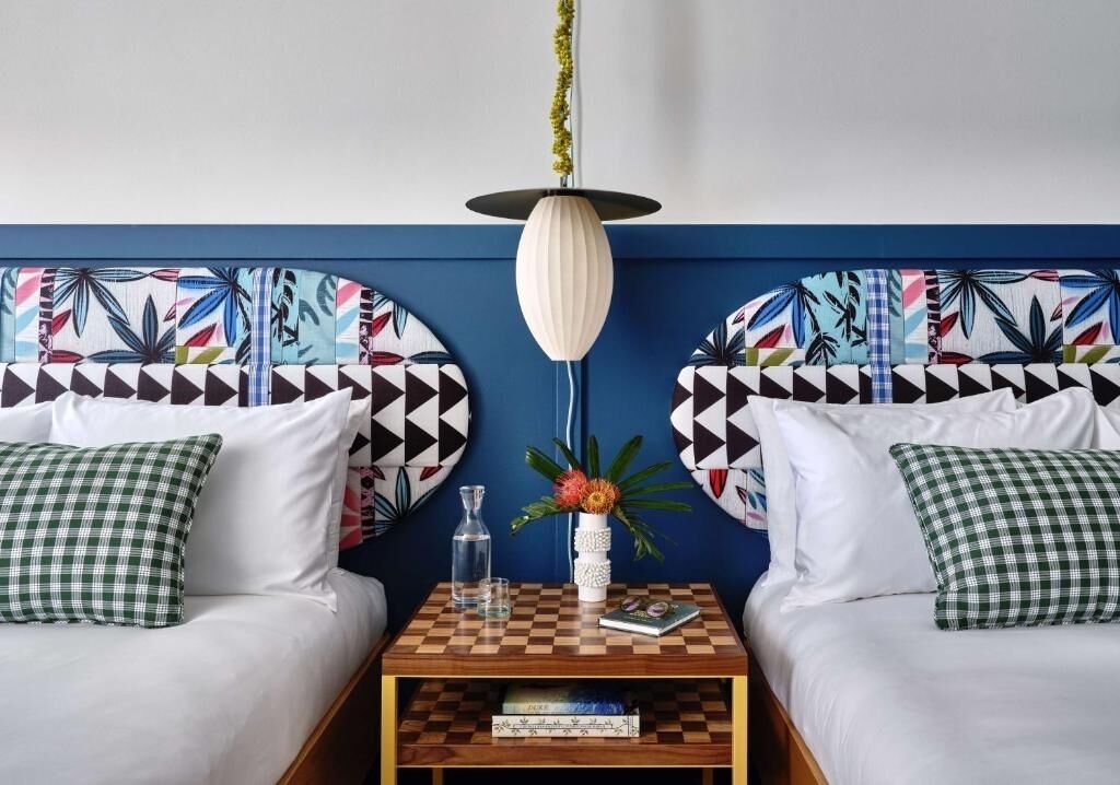 Un dormitorio con dos camas, una mesita de noche, una lámpara y una pared con estampado de hojas tropicales.