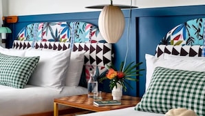 Una habitación con cabecera azul, sofá blanco, almohadas a cuadros, una mesa de centro de madera y un sombrero en el suelo.