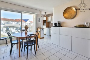 A modern kitchen with white cabinets, a dining area with a round table and chairs, and a large window offering a view of buildings. - Soleil Levant   Plage à 150m, balcon, quartier des Présidents (Les Sables-d´Olonne)