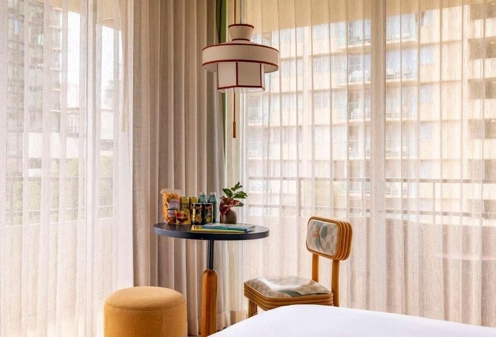 Camera d'albergo con un letto, un tavolino rotondo con una lampada, una sedia e vista attraverso tende leggere.