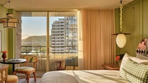 A hotel room with a bed, a table, chairs, a balcony with a view of buildings, and a hanging light fixture. - Wayfinder Waikiki | Corner King Lanai | Design Hotel, Saltwater Pool (Honolulu County)