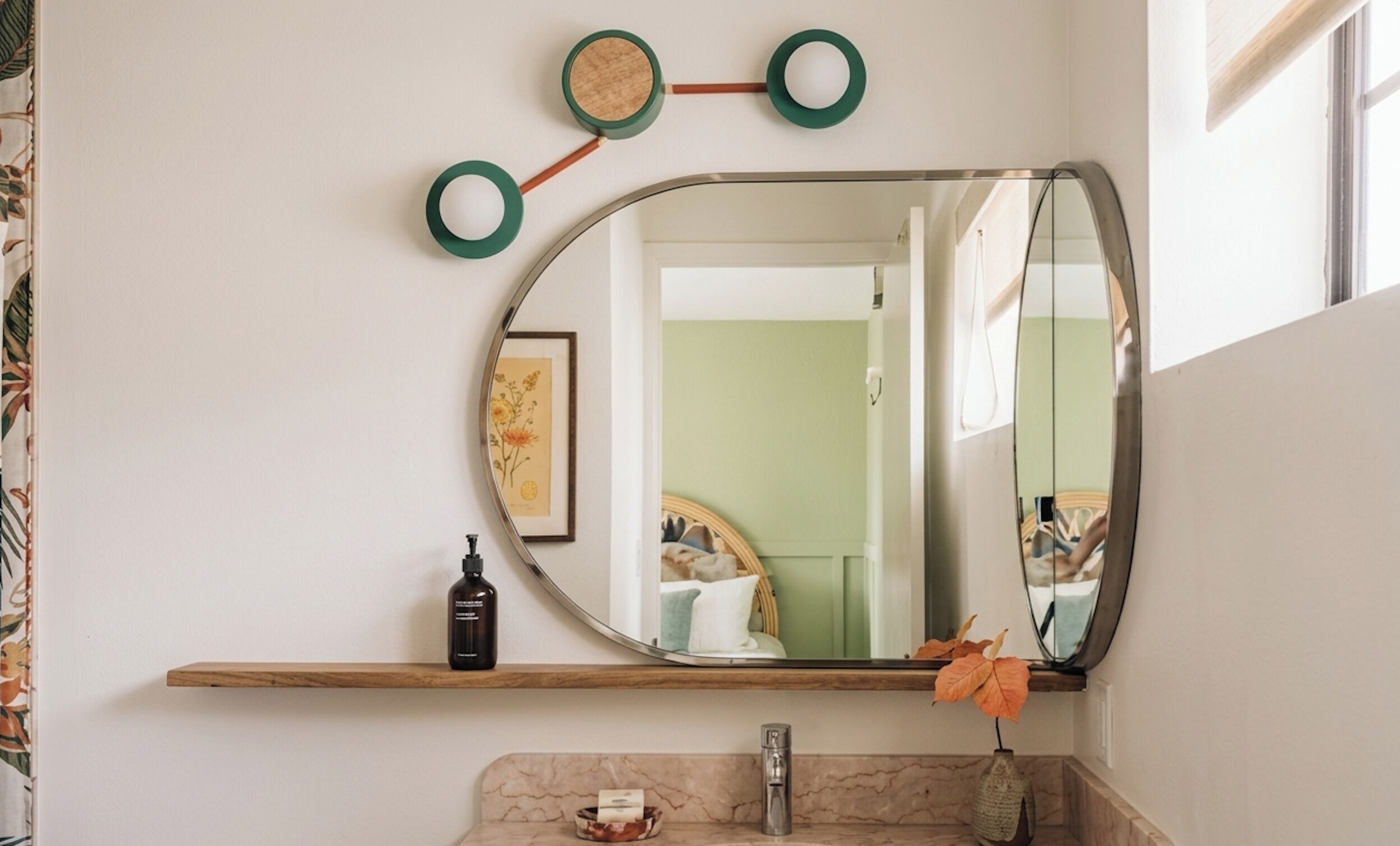 A bathroom with a large oval mirror, a wooden shelf, a marble sink, and a vase with a single leaf.