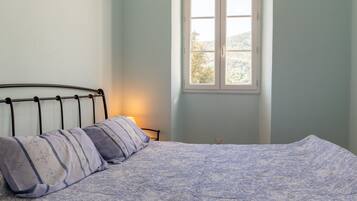 A bed with a patterned bedspread and pillows, a bedside lamp, and a window with a view of trees.