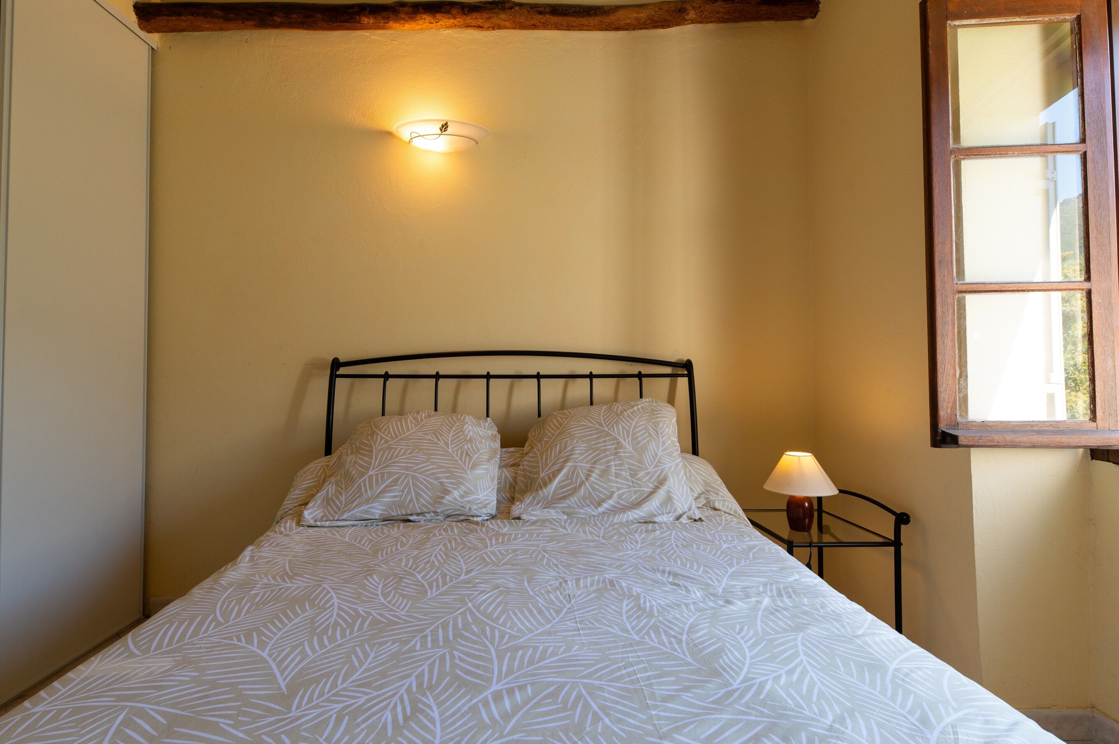 Uma cama com colcha estampada em folhas, um abajur e um ponto de luz fixado na parede.