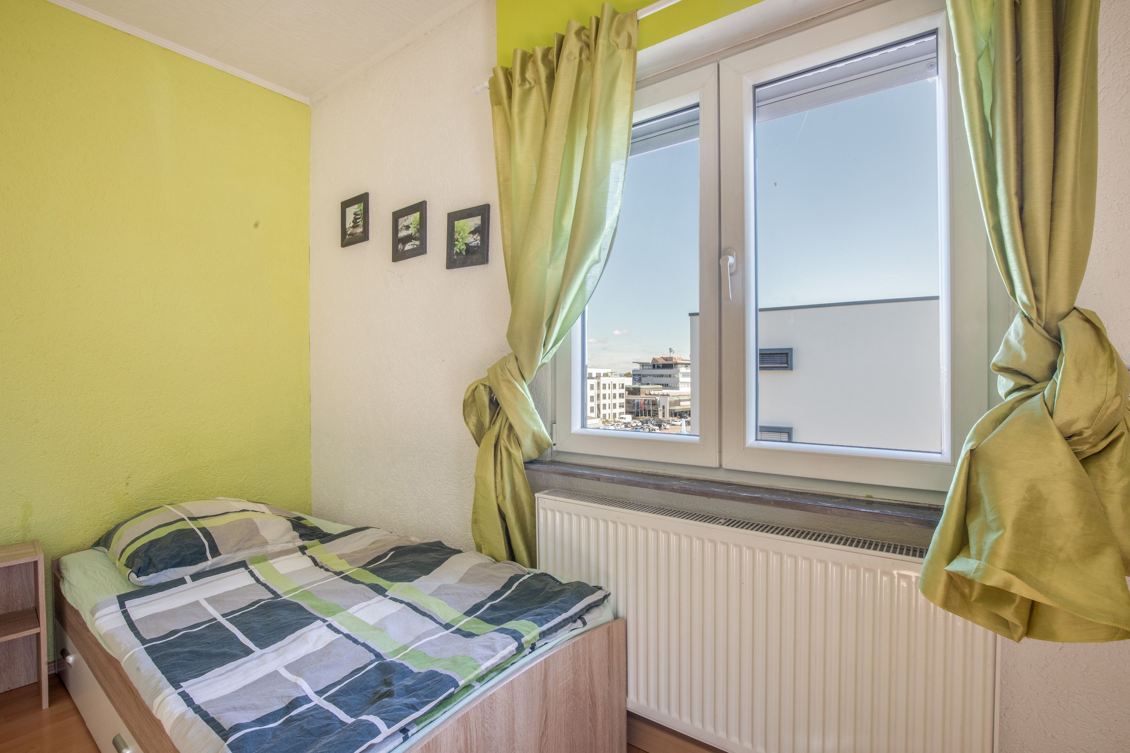 A bedroom with a bed, a window with curtains, a radiator, and a view of buildings outside.