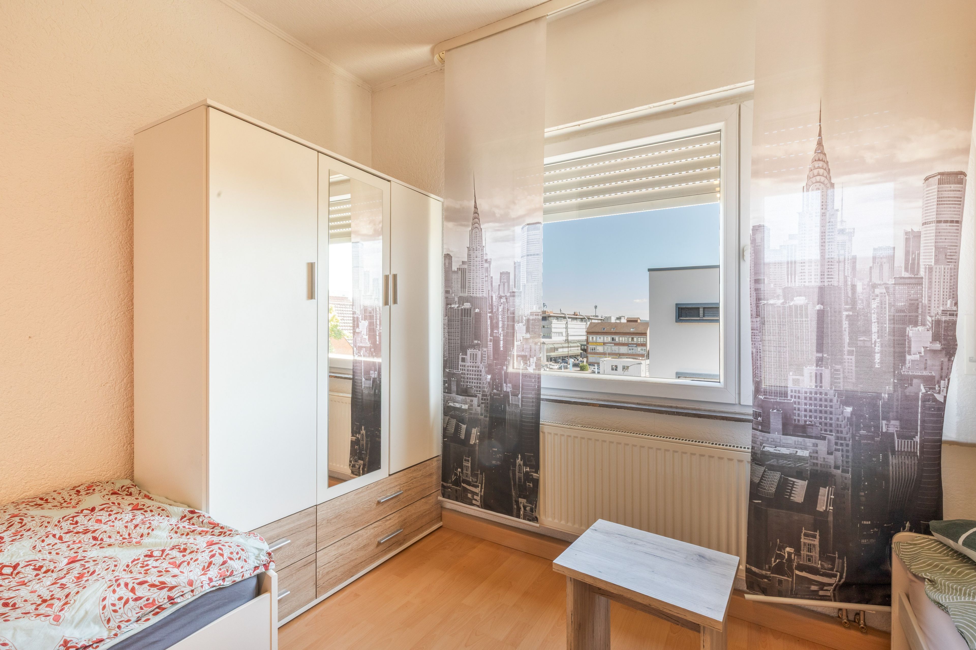 Een slaapkamer met een bed, een kledingkast, een raam met zonwering en uitzicht op een stadsgezicht.