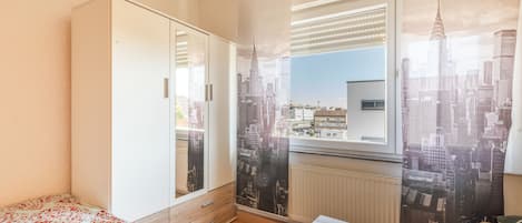 Une chambre à coucher avec un lit, une armoire, une fenêtre avec des stores et une vue sur le paysage urbain.