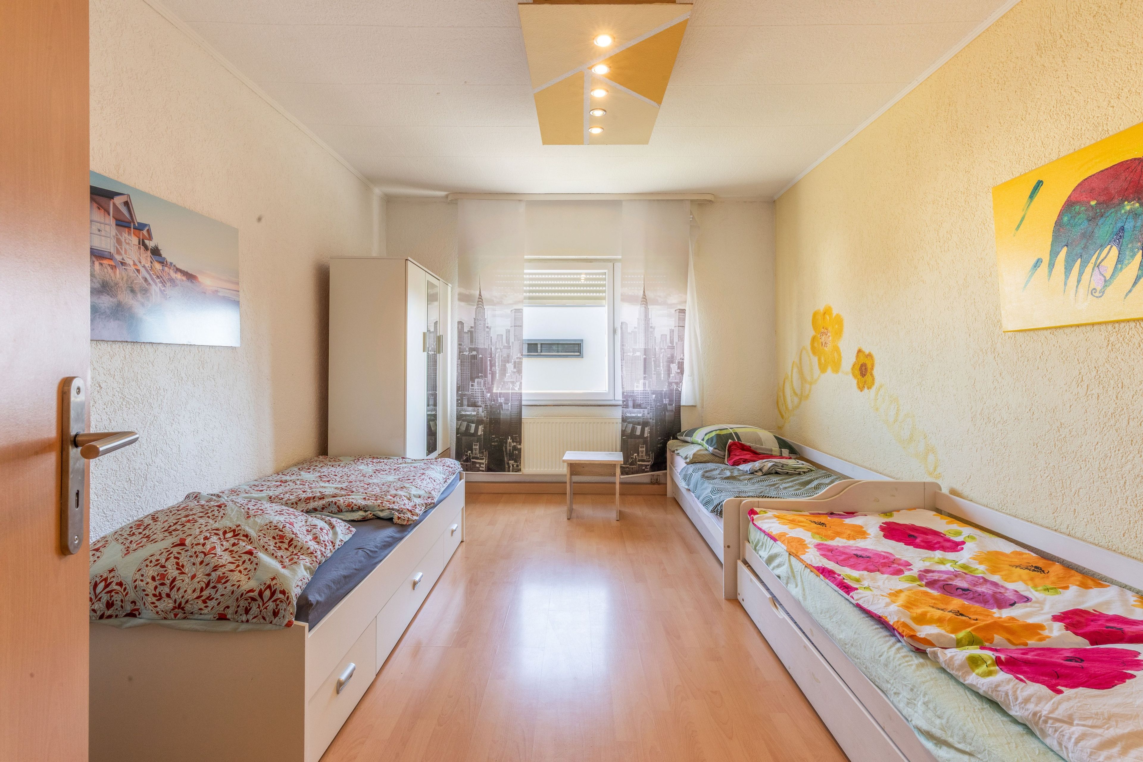 Une chambre avec deux lits simples, une petite table et une vue sur le paysage urbain par la fenêtre.