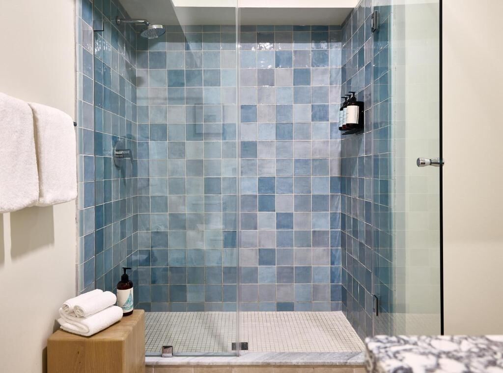 Ducha con paredes de azulejos azules, un banco de madera con toallas y puerta de cristal.