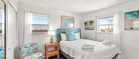 A bedroom with a bed, a chair, a nightstand, a window with a view, and a painting on the wall.