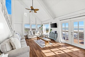 Un salon spacieux doté d’un plancher en bois, d’un ventilateur de plafond, d’un canapé, d’une table basse et offrant une vue sur la plage grâce à de grandes portes coulissantes.