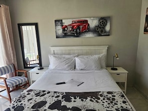 Habitación de hotel con cama, una silla, un escritorio, una lámpara y un cuadro de un coche en la pared.