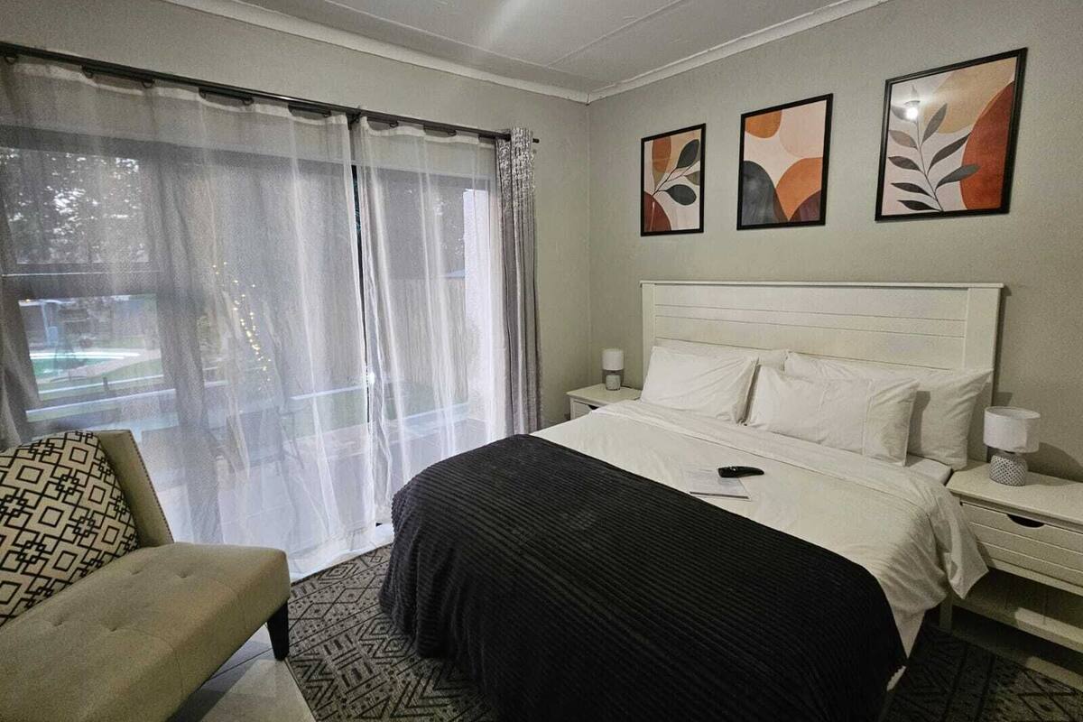 A hotel room with a bed, a chair, a nightstand, and a window with sheer curtains.