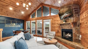 Interior acogedor de una cabaña con chimenea de piedra, un sofá grande y una zona de comedor con mesa y sillas.