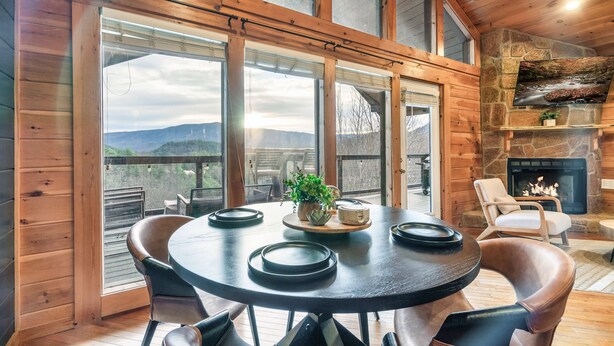 Un interior de madera con una mesa redonda, sillas, chimenea y ventanas amplias.