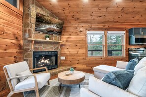 A log cabin interior with a stone fireplace, white furniture, and a view through a window. - Epic Couples Getaway! Mountain Views! Hot Tub! (Sevierville)
