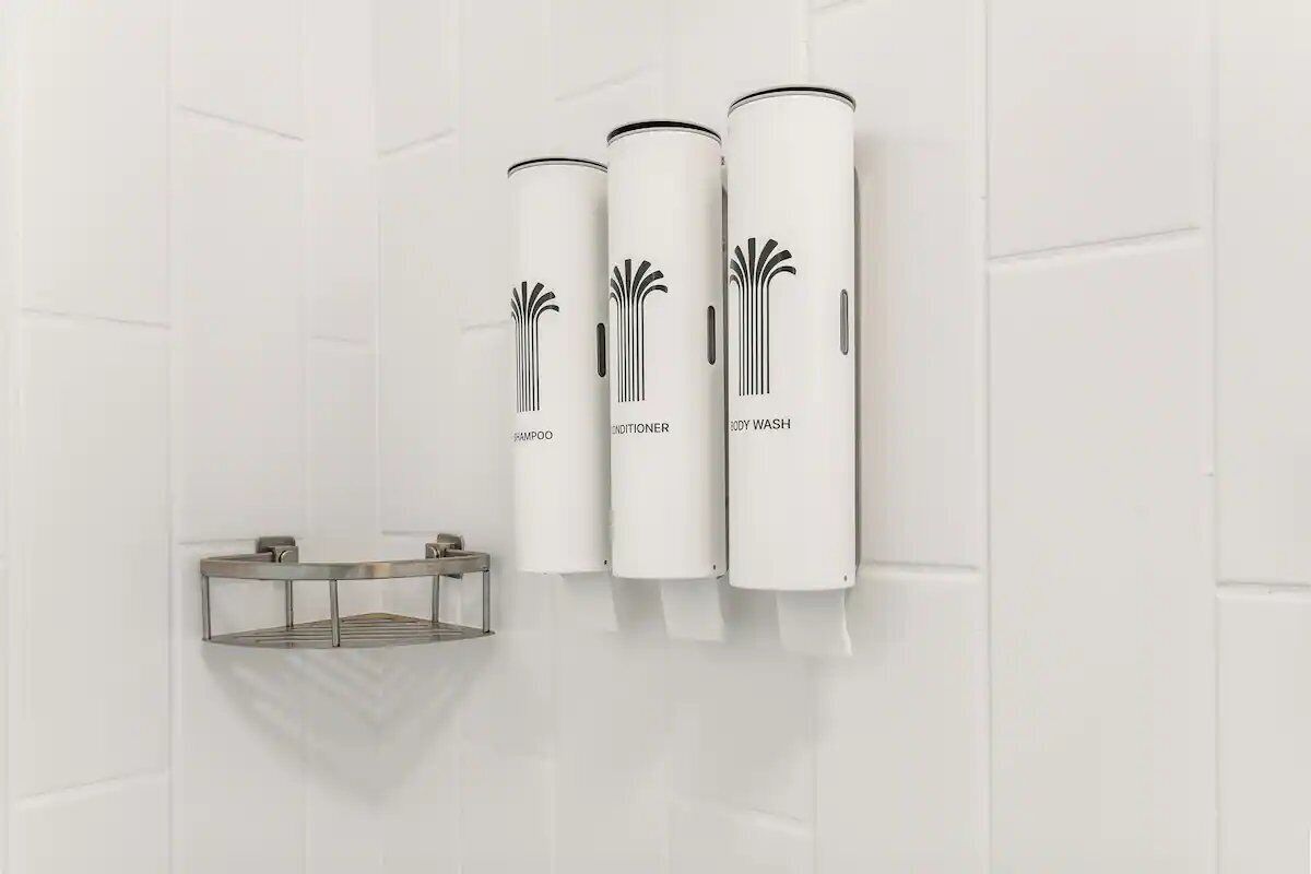 A bathroom with three cylindrical dispensers labeled 'SHAMPOO,' 'CONDITIONER,' and 'BODY WASH' mounted on a white tiled wall.