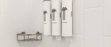 A bathroom with three cylindrical dispensers labeled 'SHAMPOO,' 'CONDITIONER,' and 'BODY WASH' mounted on a white tiled wall.