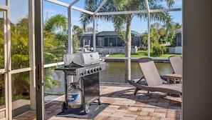 Een overdekt terras met een barbecue, lounge stoelen en uitzicht op een huis en water.