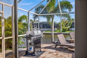 A covered outdoor area with a grill, lounge chairs, and a view of a house and water. - Triple Palms Vacation Home, With Heated Pool (Cape Coral)