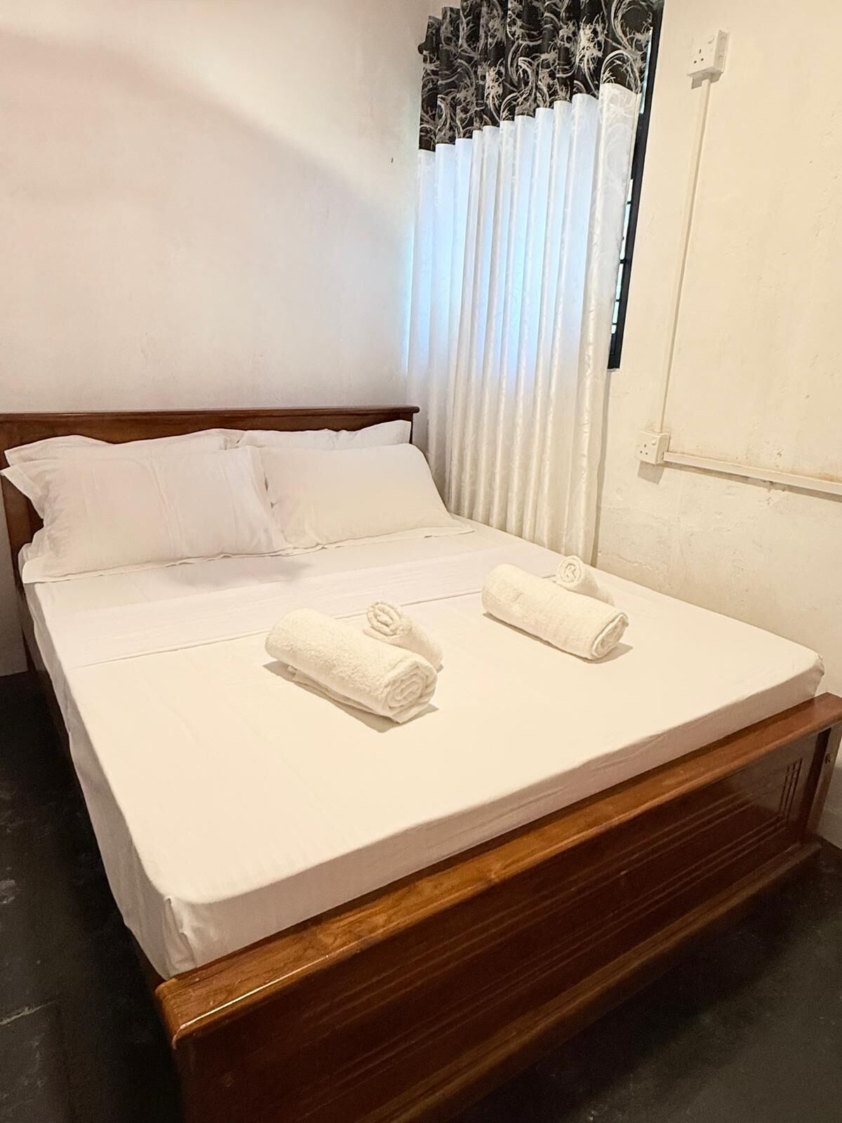 A neatly made bed with white linens and two rolled towels, a wooden headboard, and a window with curtains.