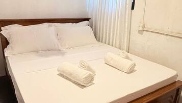 A neatly made bed with white linens and two rolled towels, a wooden headboard, and a window with curtains.