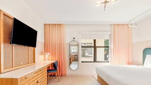 Hotellihuone, jossa on sänky, työpöytä tuolilla, televisio, peili ja ikkuna, jossa on verhot.