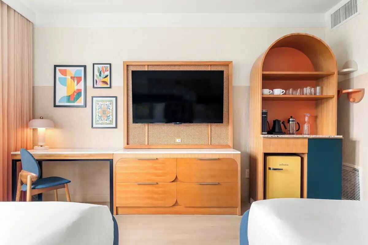 A hotel room with a desk, two beds, a television, a small refrigerator, and a kitchenette.