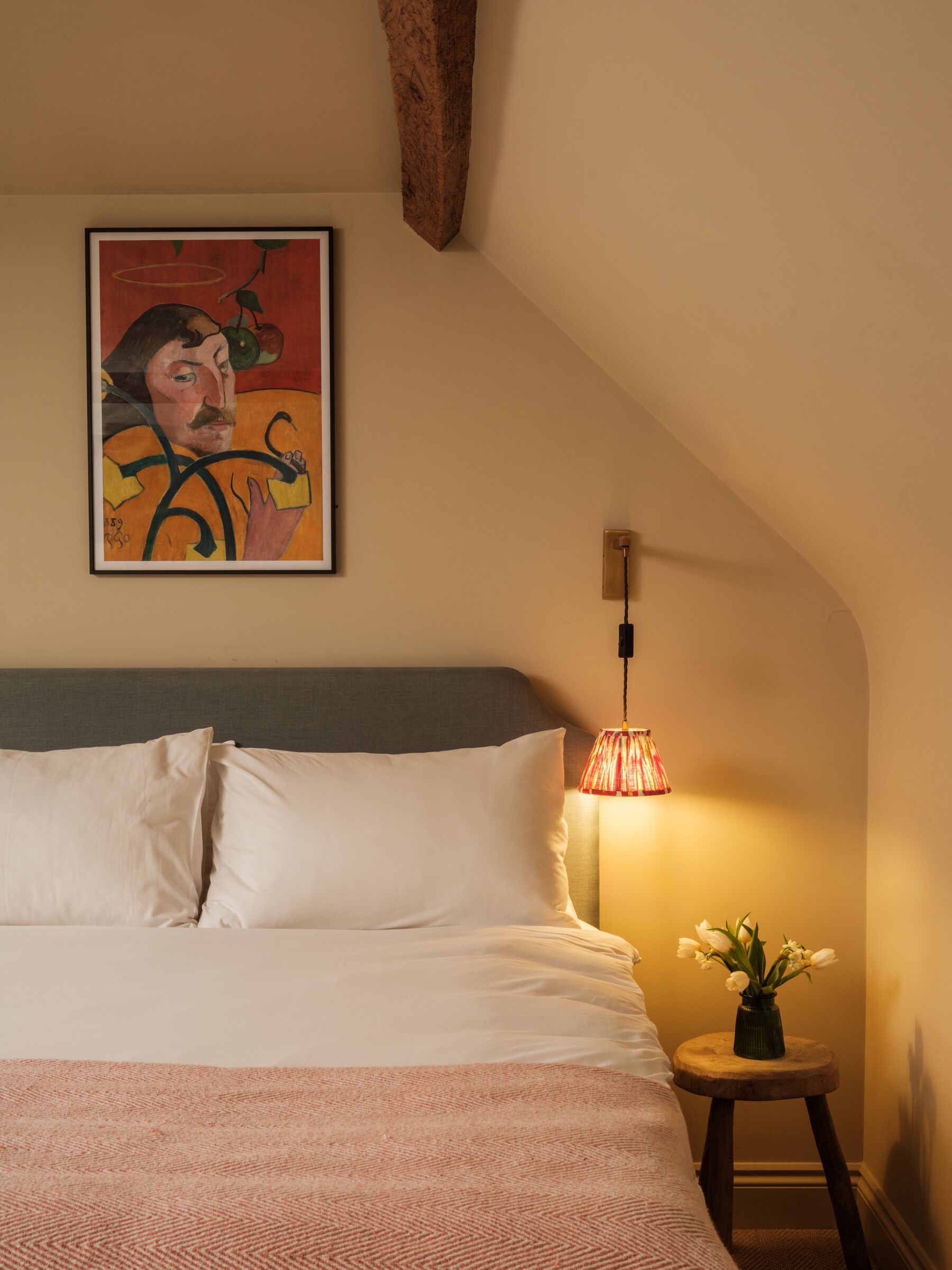 A bedroom with a bed, a lamp, a painting, and a vase with flowers.