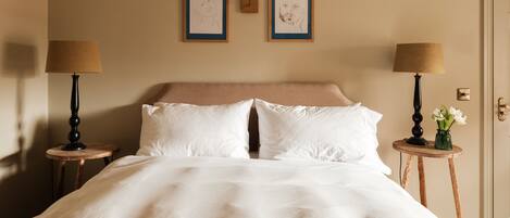 Una cama bien hecha con sábanas blancas, dos cuadros enmarcados en la pared y dos mesitas de noche con lámparas.