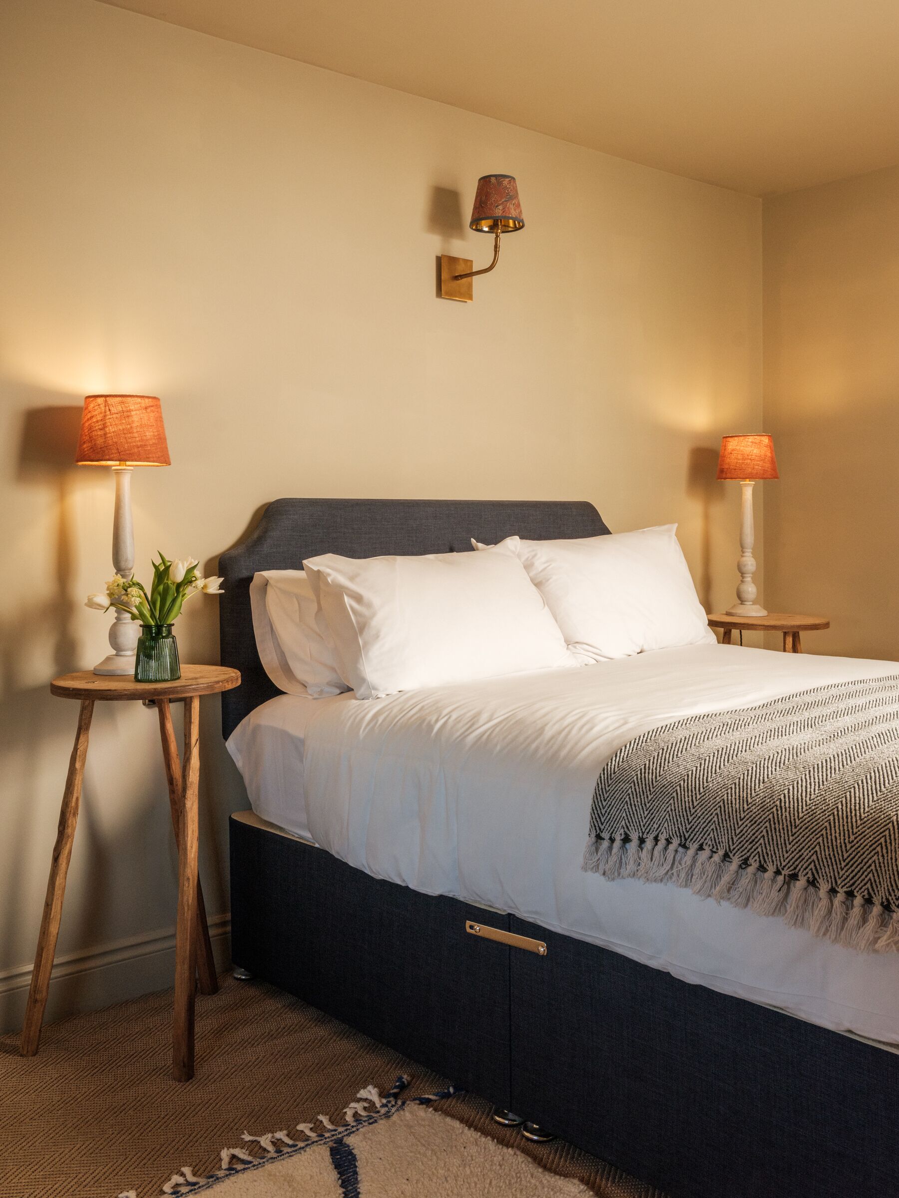 A neatly made bed with white linens, two bedside lamps, a small table with a vase of flowers, and a wall-mounted lamp.