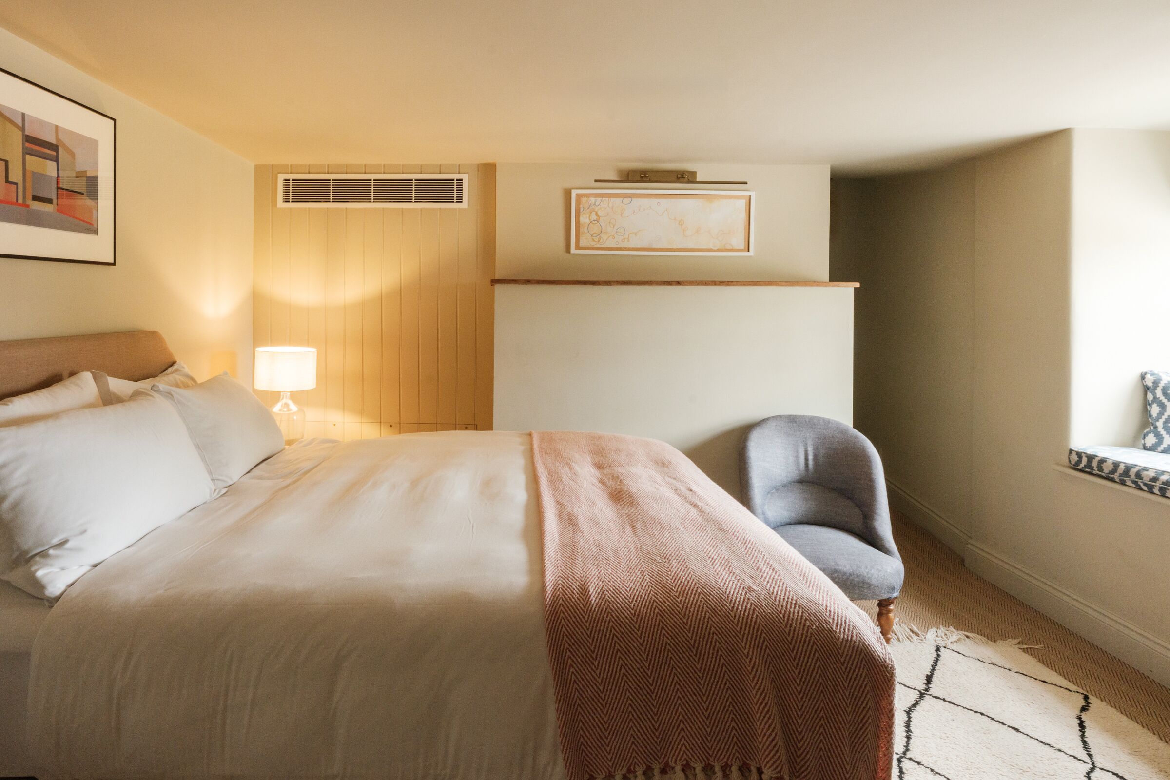 Een hotelkamer met een bed, een stoel, een lamp, een kunstwerk aan de muur en een raam met uitzicht.