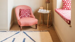 A cozy room with a pink armchair, a small table with a lamp, a framed picture on the wall, and a window with a red patterned cushion. - The Bell & Crown (Zeals)