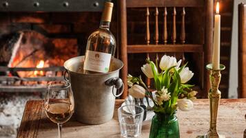 A dining table with a bottle of wine, a candle, a vase with flowers, and a fireplace in the background.