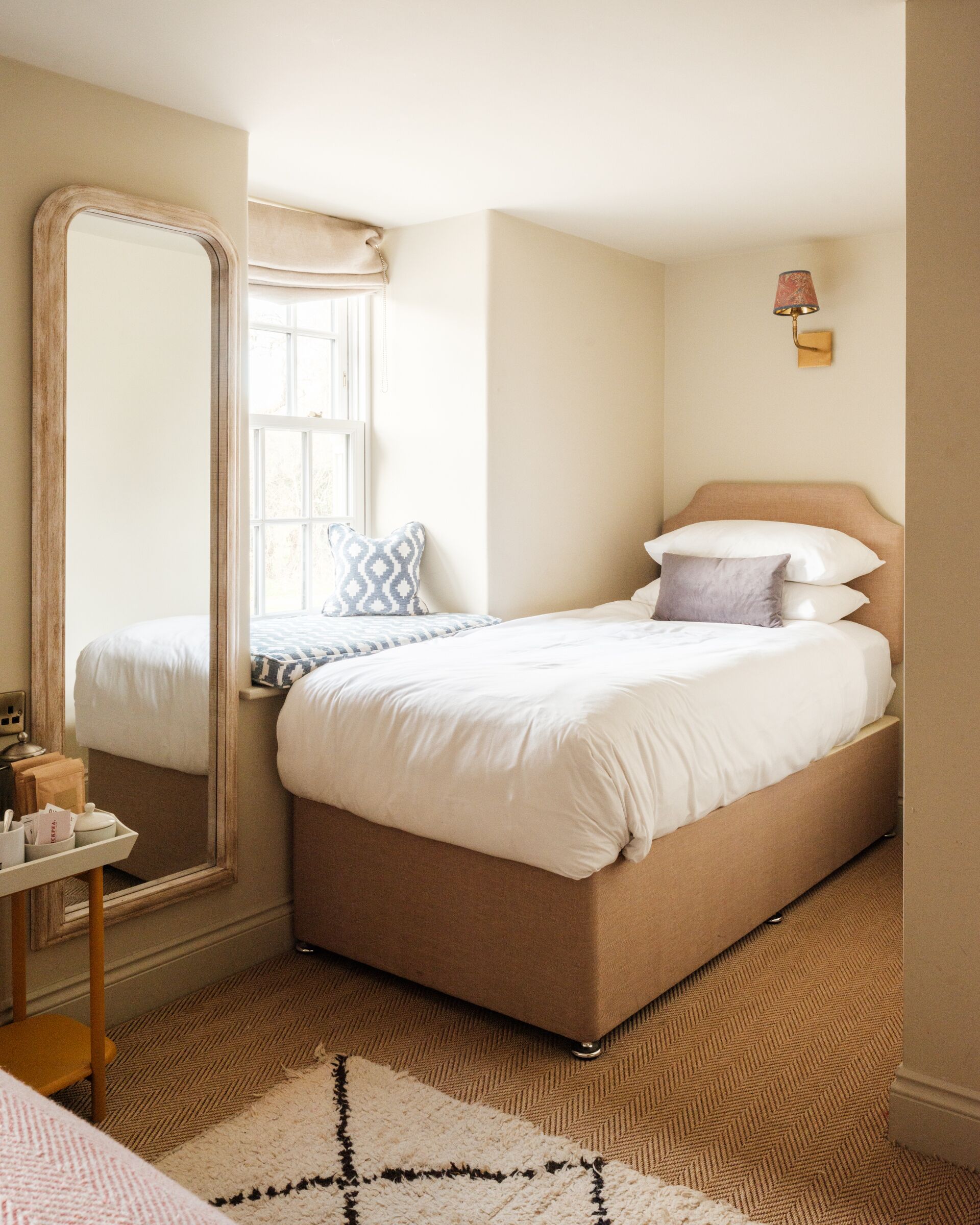 A hotel room with a large mirror, a bed with white bedding, a patterned rug, and a small table with a lamp.