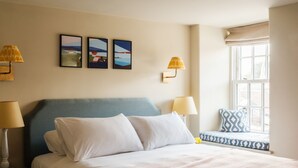 A bedroom with a bed, two wall-mounted lamps, a window with a view, and three framed pictures on the wall. - The Bell & Crown (Zeals)